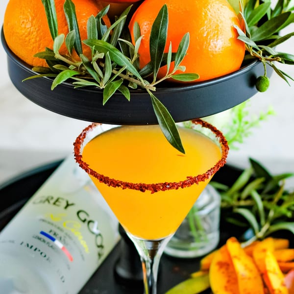 Orange cocktail with a chili-lime rim, Grey Goose bottle, and oranges with olive branches.