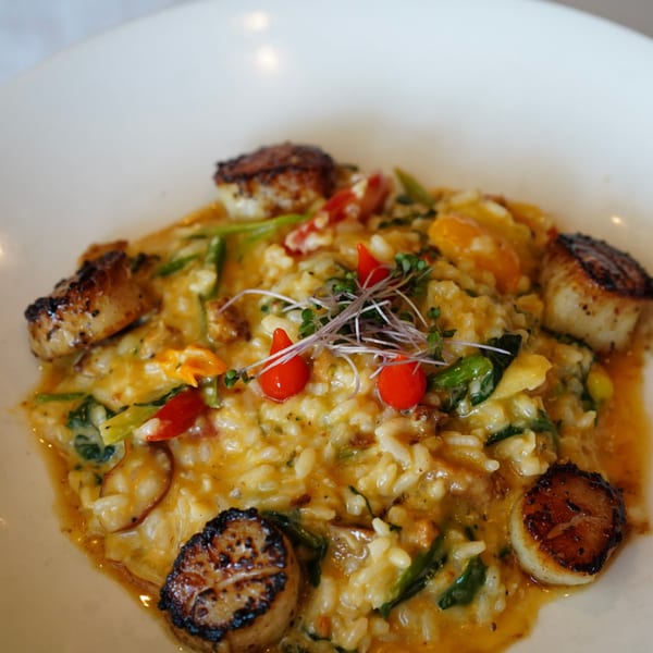 Seared scallops on a bed of creamy risotto with red peppers and microgreens in a white bowl.