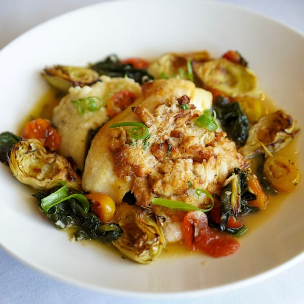 White fish with crab meat, mashed potatoes, roasted artichokes, tomatoes, and greens in a white bowl.