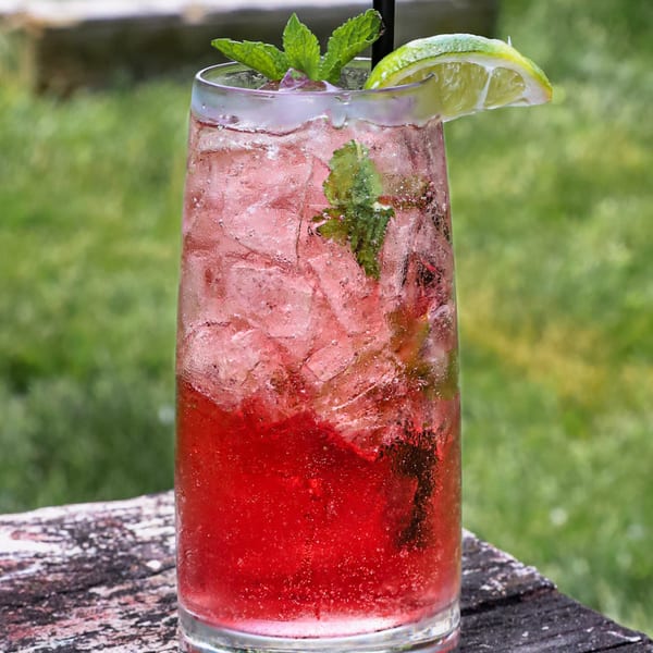 A tall glass of red iced drink with mint leaves and a lime wedge on a wooden surface.