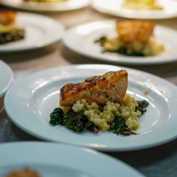 Plated salmon with couscous and sautéed greens, ready to be served in a restaurant kitchen.