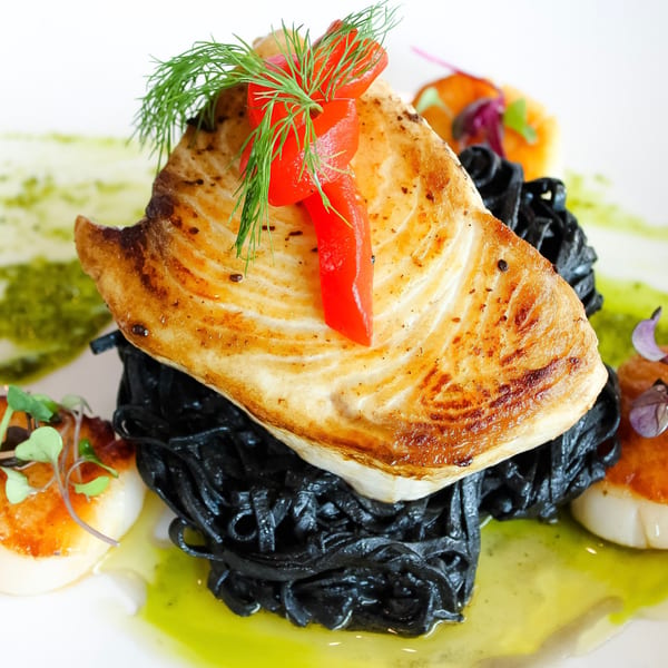 Grilled fish with red peppers and dill on black pasta, surrounded by scallops and green sauce.