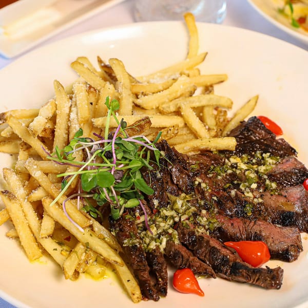 Sliced steak with chimichurri sauce, french fries, microgreens, and red peppers on a white plate.